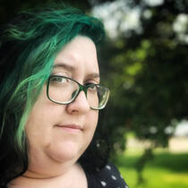 This image is a picture of Maery. She is a white person in her thirties with glasses and black and green hair.